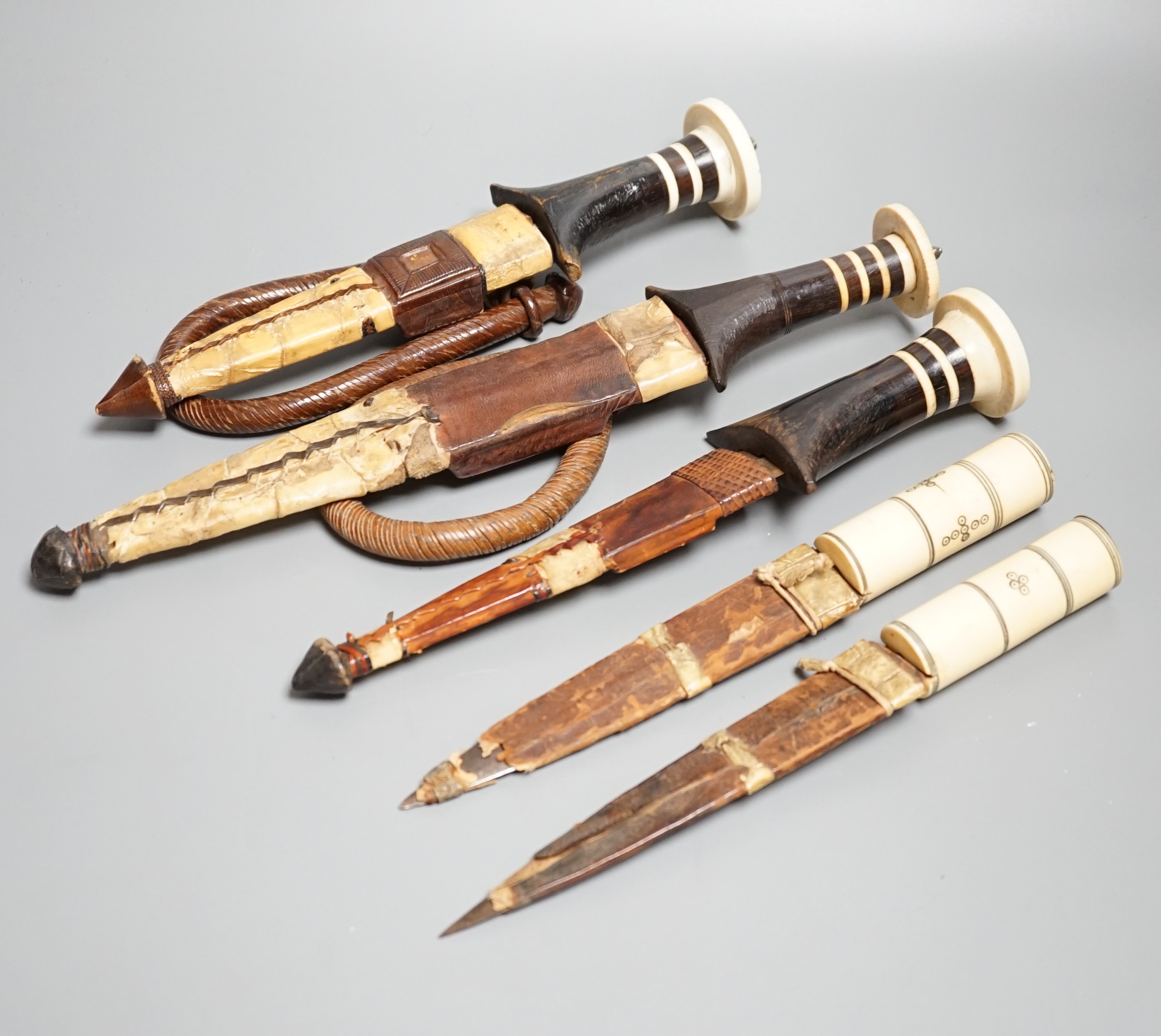 Five assorted African Nilotic tribal daggers c.1900, comprising three with turned ivory mounted ebony handles (two with silver pommels); and two further with cylindrical ivory handles, all in their leather sheaths, maxim